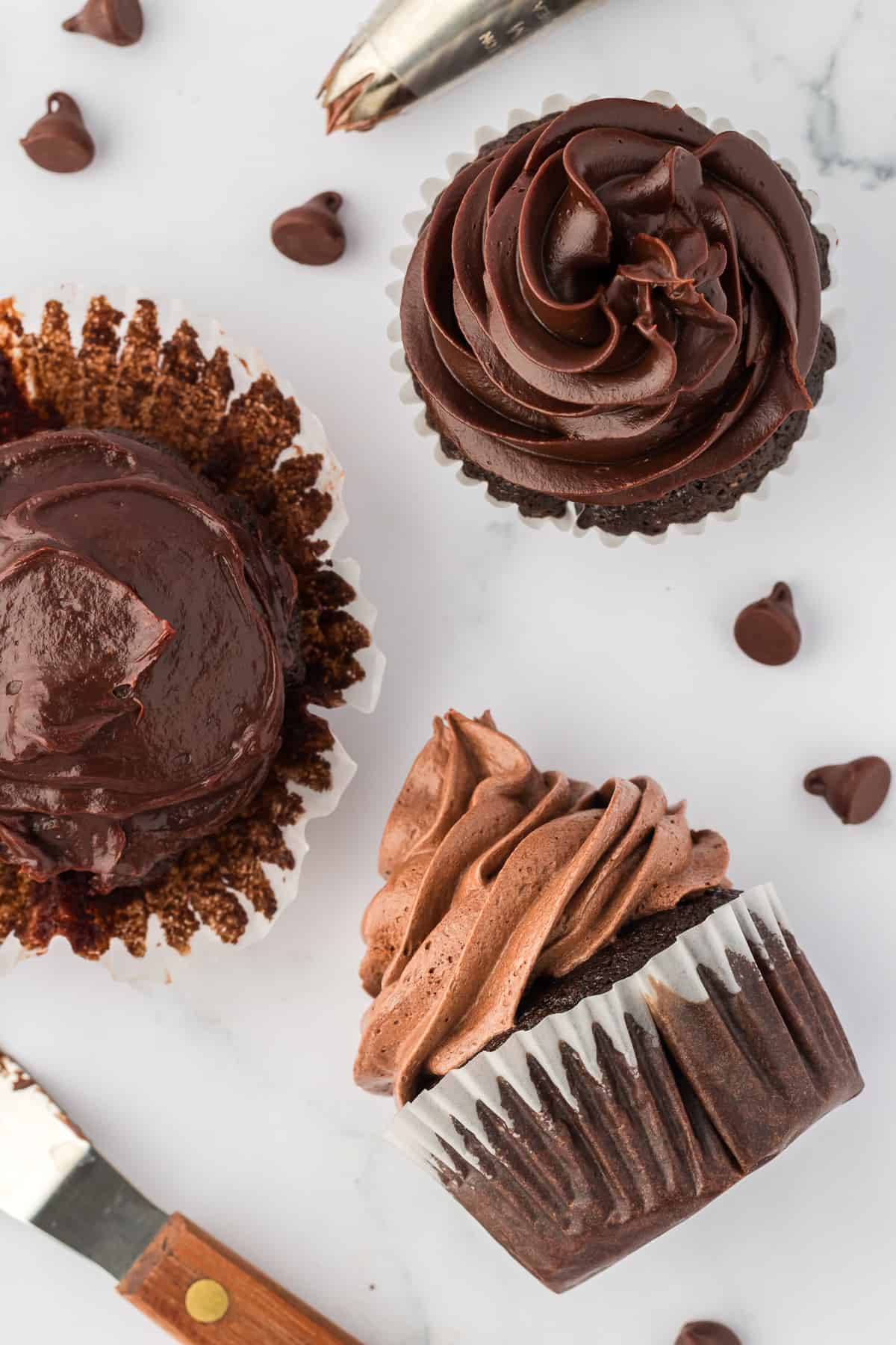 three chocolate cupcakes with chocolate chips sprinkled around. The one with regular ganache with its paper liner peeled partially off, the piped ganache cupcake sitting in a paper liner and the cupcake with whipped ganache laying on its side