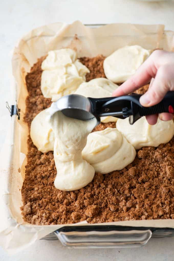 coffee cake batter being dropped into a cake pan with parchment paper with an ice cream scoop over crumb topping 