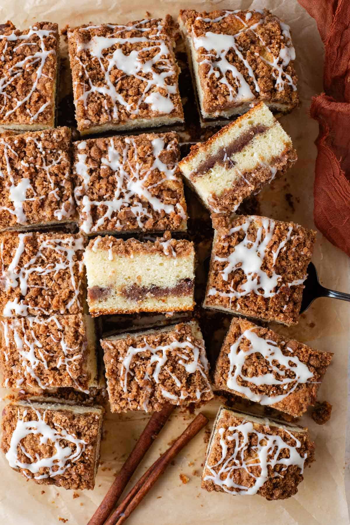 coffee cake slices arranged on brown paper with some on their sides, a fork, orange towel and cinnamon sticks