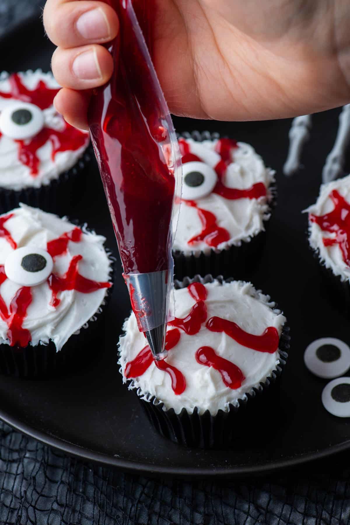 piping red bloody jam onto an eyeball cupcake with more eyeball cupcakes around it