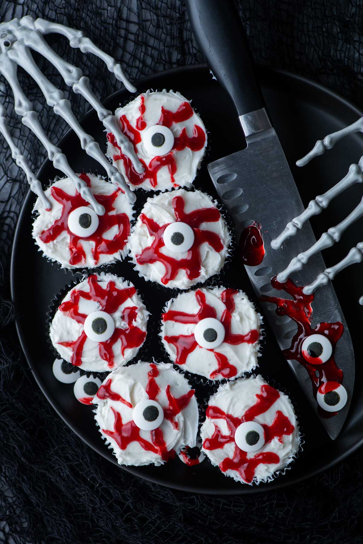 eyeball cupcakes on a black plate with a large knife covered in bloody jam, more candy eyeballs and skeleton hands