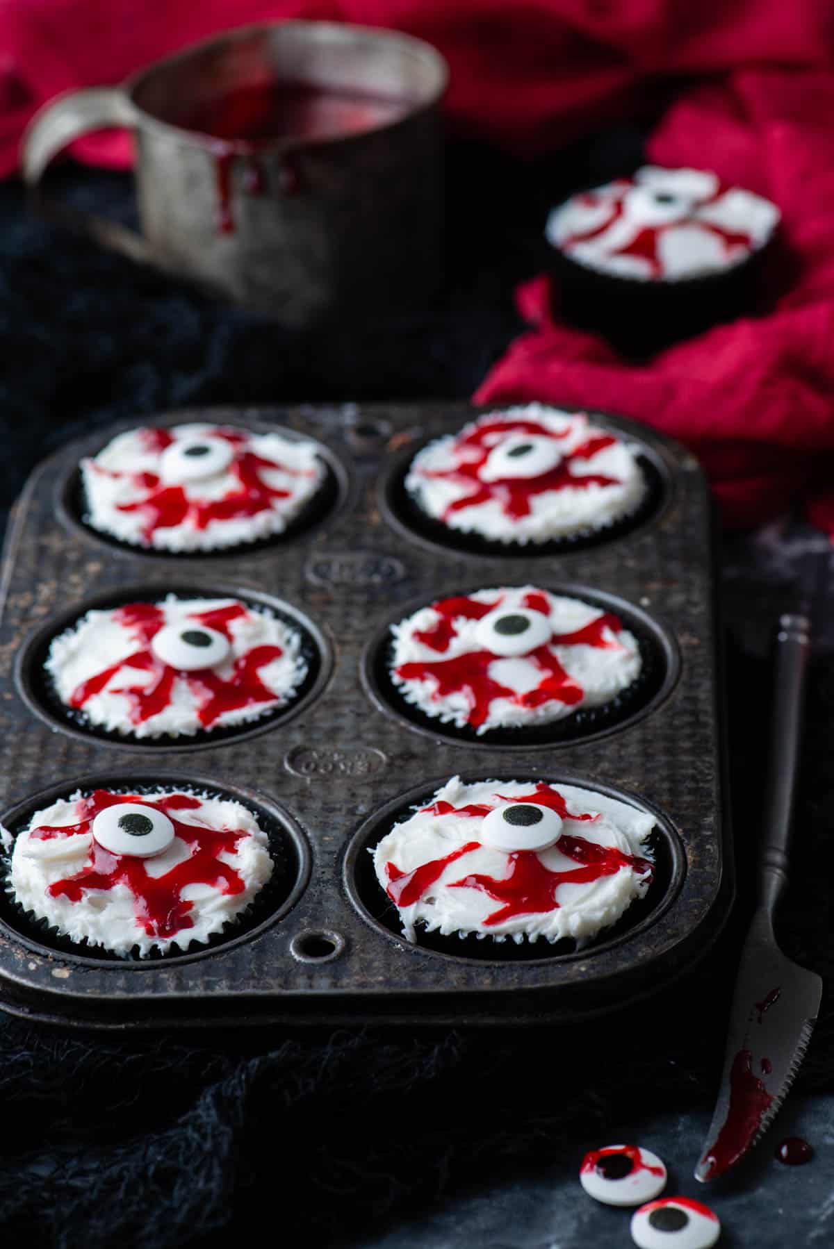 a cupcake tin full of eyeball cupcakes surrounded by a knife, candy eyeballs, bloody jam and a red towel