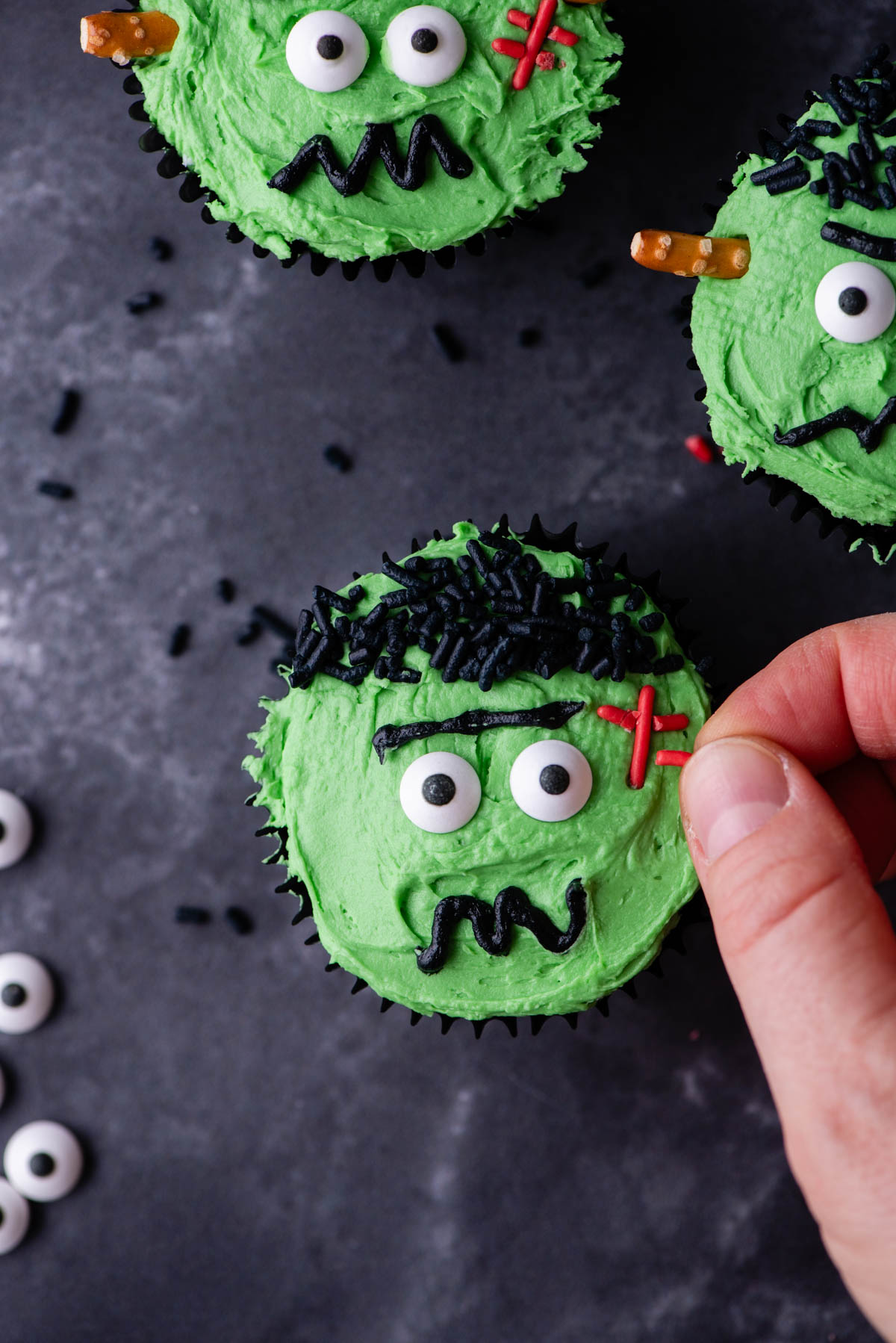 A frankenstein cupcake being decorated by adding red sprinkles for the stitch with more completed frankenstein cupcakes near it