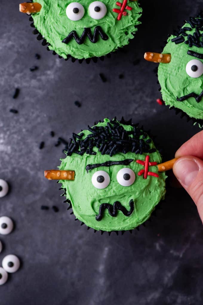 A frankenstein cupcake being decorated by adding pretzel sticks for the neck bolt with more completed frankenstein cupcakes near it