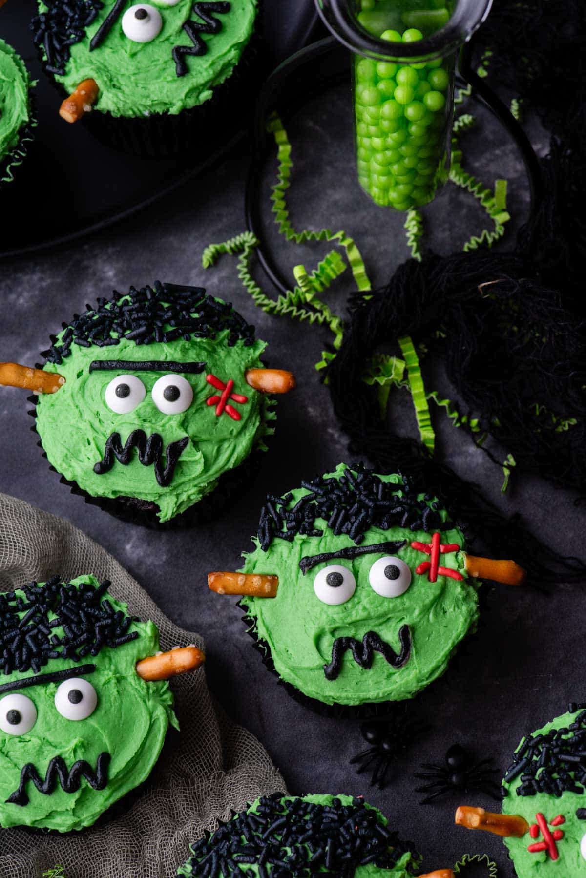 frankenstein cupcakes on a grey surface with green sprinkles and tiny streamers beside them