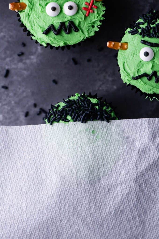 frankenstein cupcakes near another cupcake partially covered with a paper towel with black sprinkles on the uncovered part