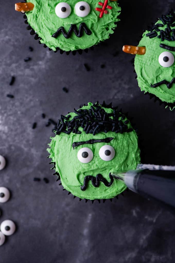 a frankenstein cupcake being decorated by piping on a mouth, sitting beside more completed frankenstein cupcakes