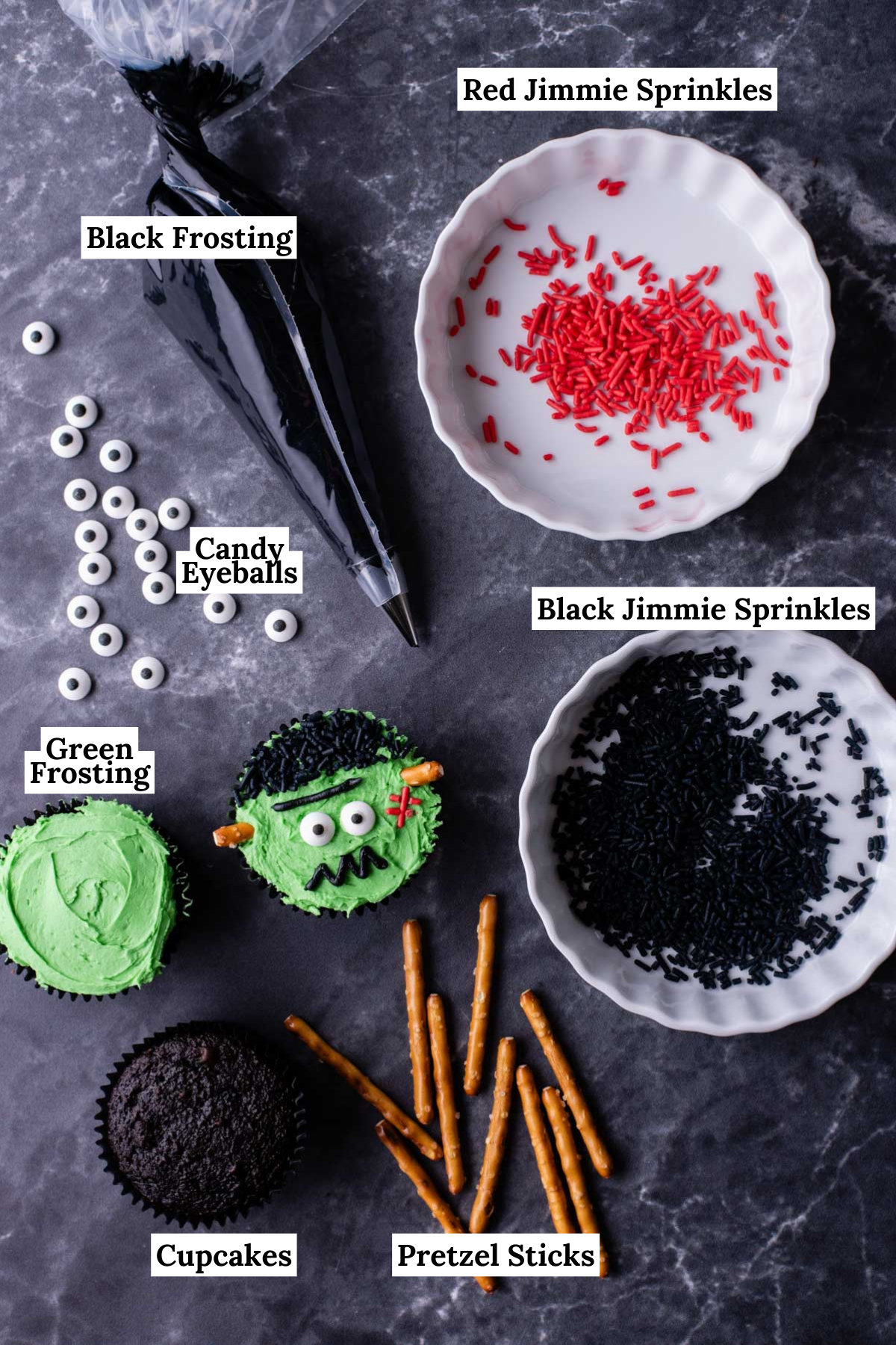 ingredients for Frankenstein cupcakes including red sprinkles, black sprinkles, chocolate cupcakes, pretzel sticks, candy eyeballs, and black frosting