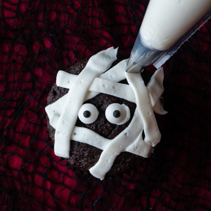 piping white strips of frosting to create a mummy look on a chocolate cupcake sitting on a red and black surface with candy eyes in the middle of it