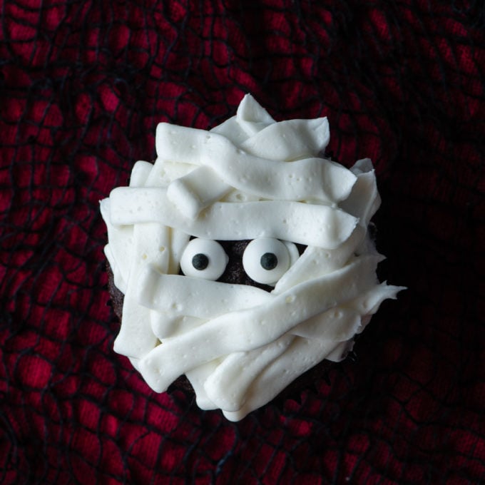 a cupcake decorated as a mummy with candy eyeballs and strips of white frosting on a red and black surface