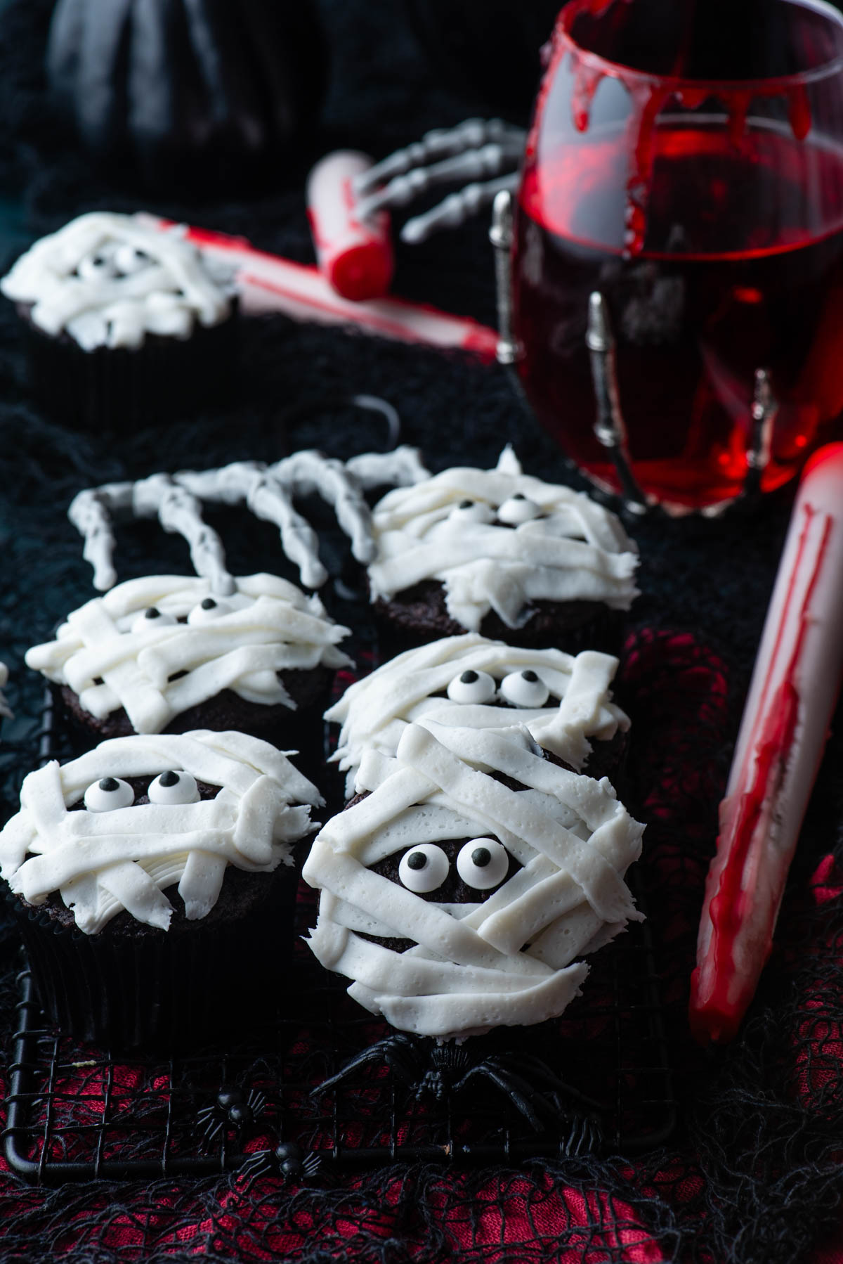 mummy cupcakes on a black and red surface with a glass of red liquid, a skeleton hand and little black plastic spiders scattered around