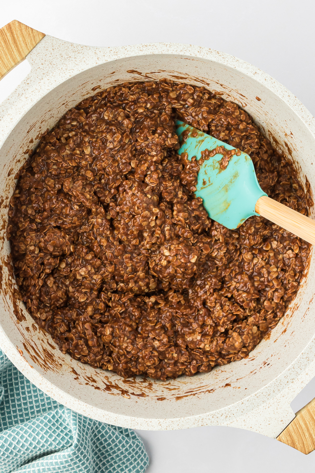 No bake cookies mixture in a white pot with a teal spatula stick into it and a teal kitchen towel beside it