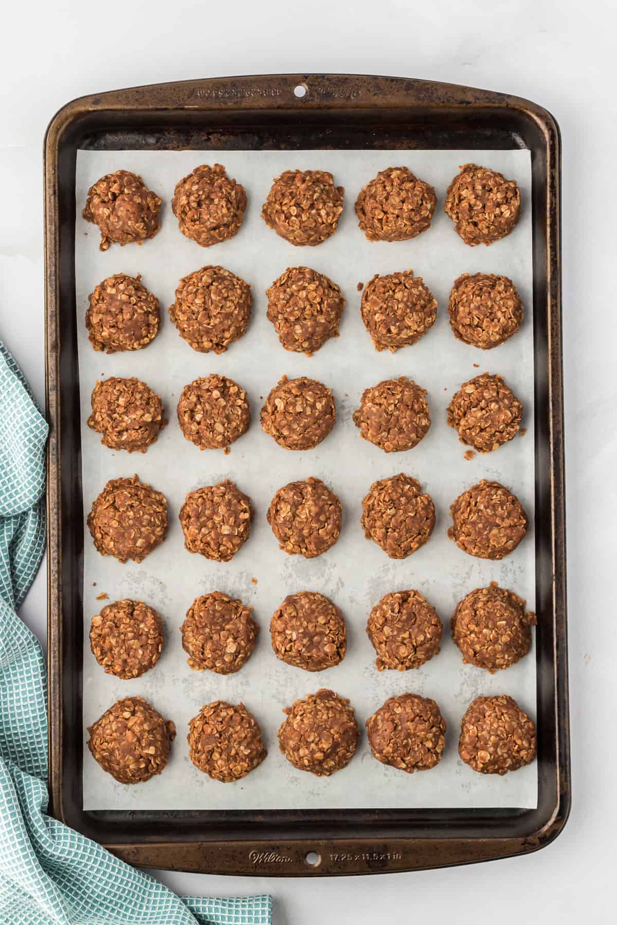 no bake cookies lined up filling up a baking sheet lined with parchment paper with a blue kitchen towel to the left of it