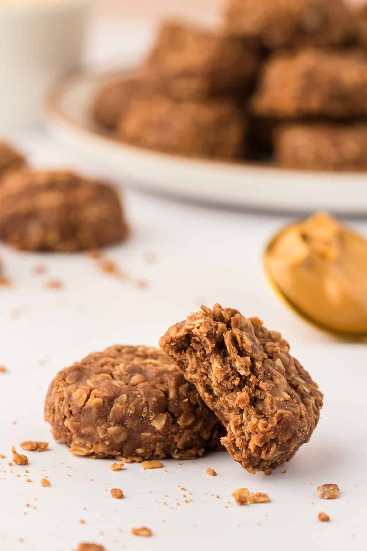 two no bake cookies, one with a bite out leaning on top of the other one, with more cookies and a spoon full of peanut butter in the background