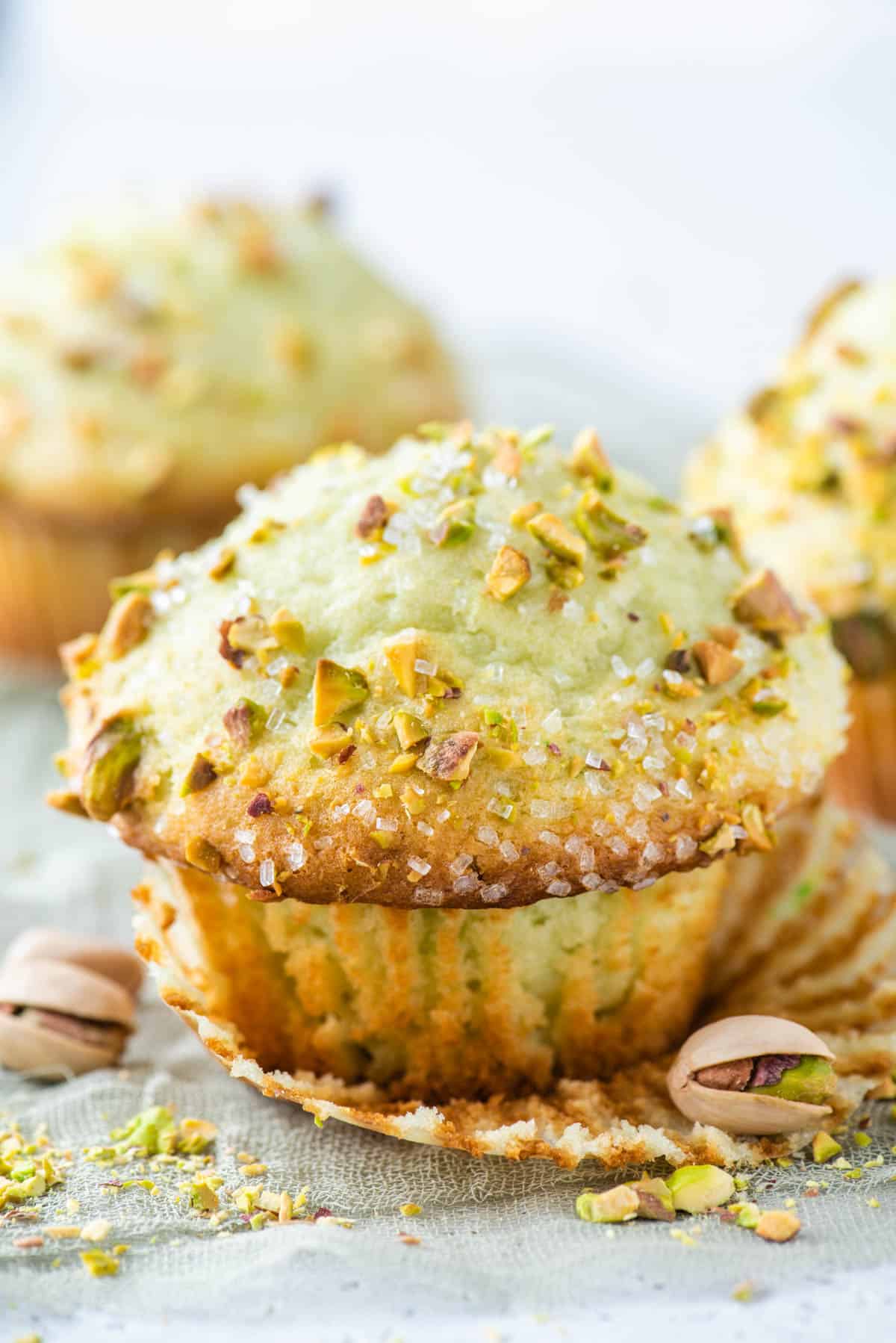 a pistachio muffin with its muffin paper pulled off with whole pistachios around it and more muffins in the background