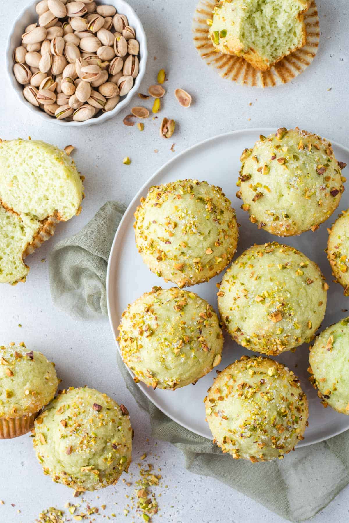 overhead view of a plate full of pistachio muffins with more muffins to the left of it, one cut in half, and a bowl of whole pistachios