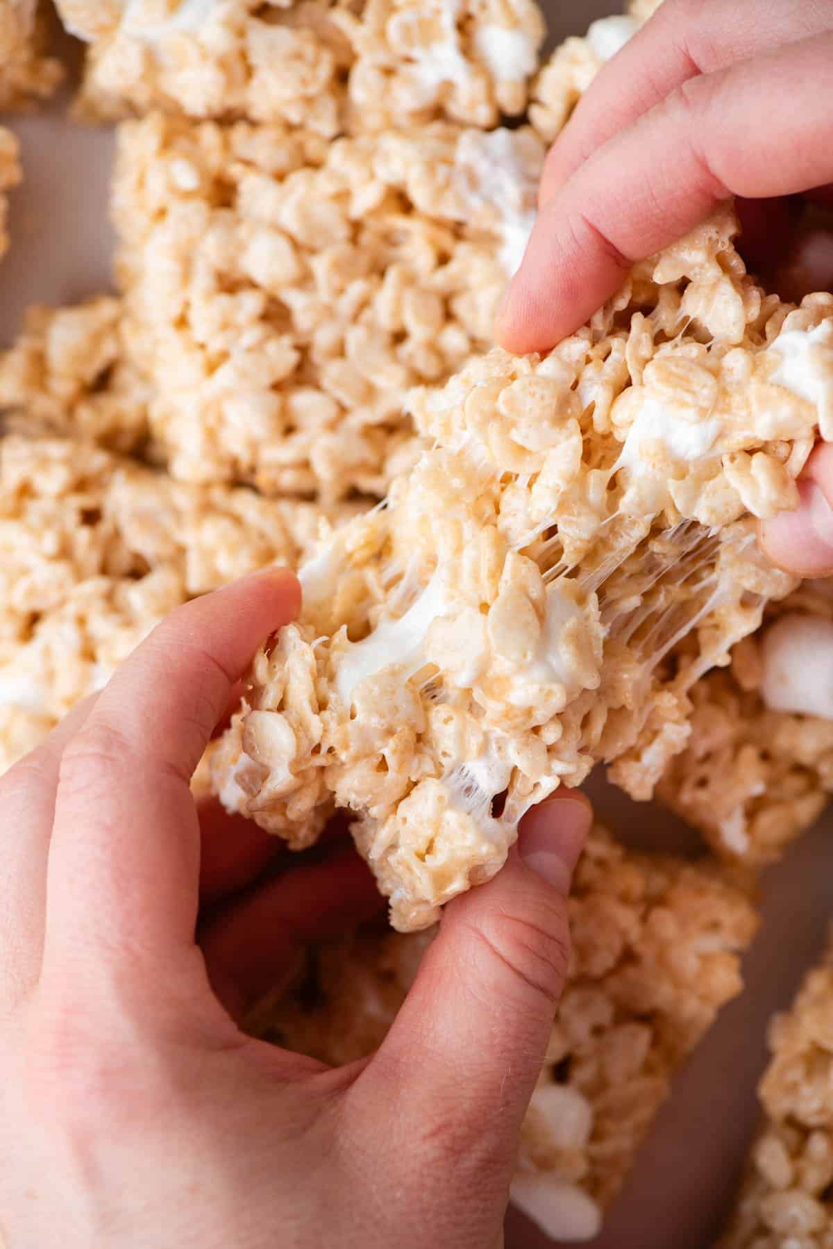 two hands pulling apart a gooey rice krispie treat over top of a pile of more rice krispie treats