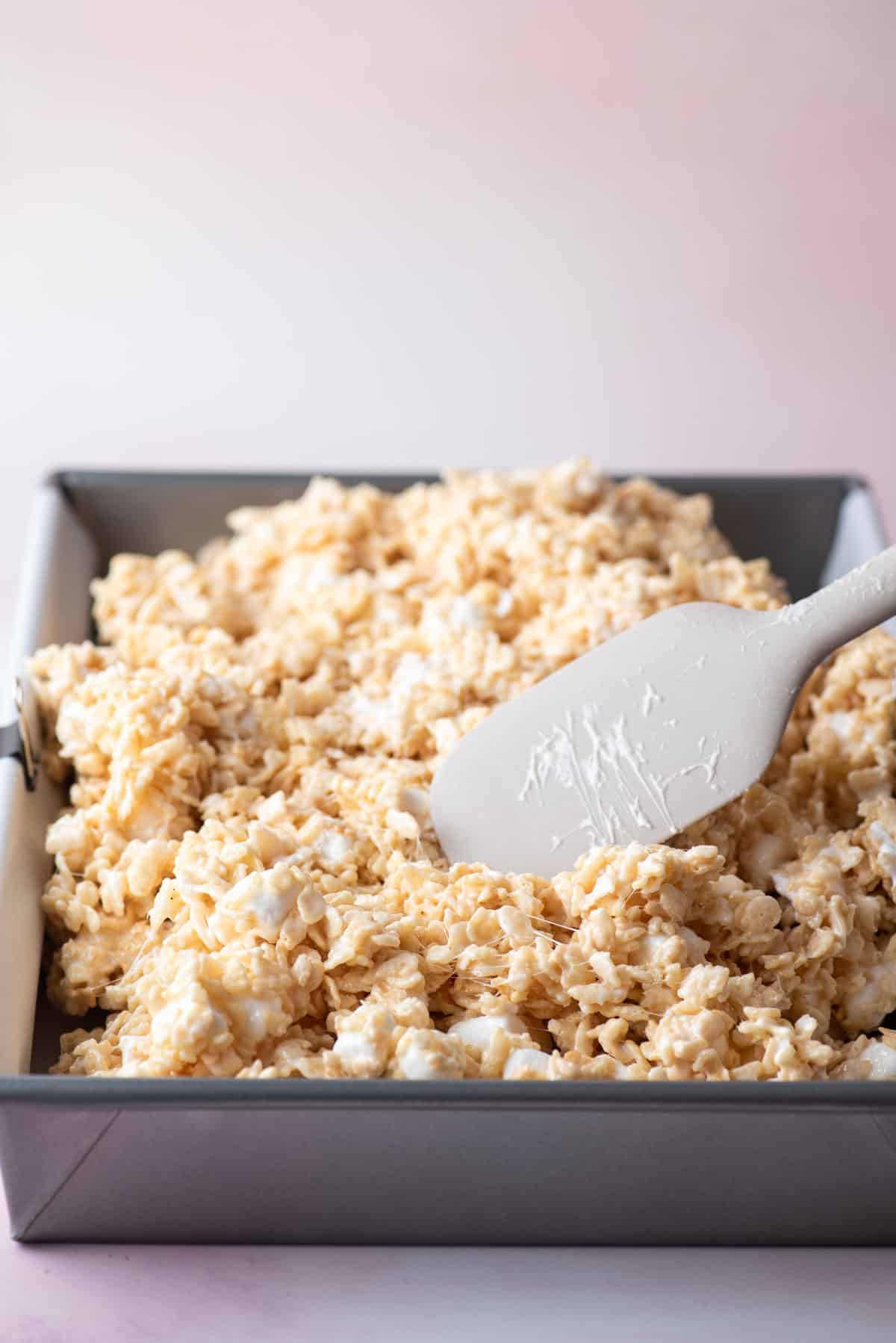 rice krispie treats mixture being gently pressed into a baking pan with a grey spatula
