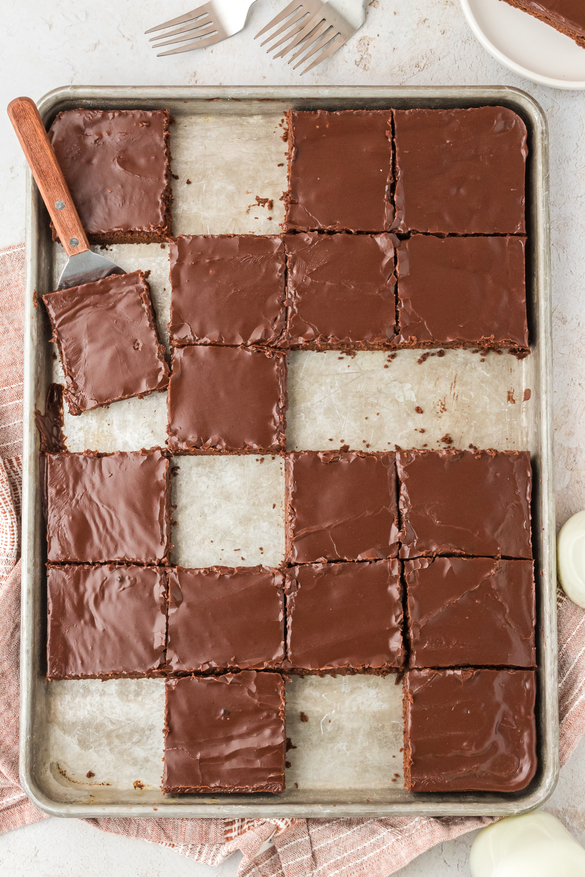 square slices of texas sheet cake on a large sheet pan with some squares missing, a spatula under one slice, a few forks and a plate with a slice of cake beside it