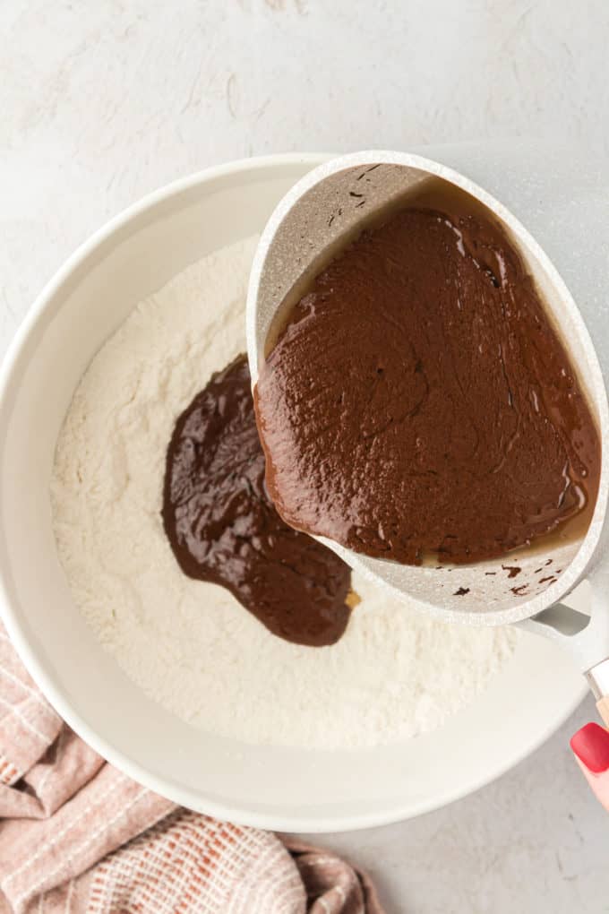 a white bowl with a flour mixture in it and the warm chocolate wet ingredients being poured into the bowl from a saucepan, and a tan towel in the left corner