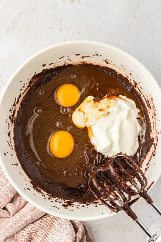 texas sheet cake batter in a white bowl with two raw eggs and sour cream on top, mixers covered in chocolate batter leaning on the bowl and a tan kitchen towel to the left of the bowl