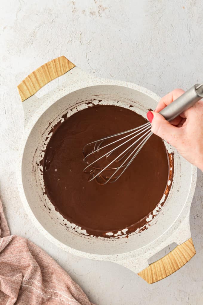 chocolate frosting in a saucepan being whisked with a wire whisk and a tan kitchen towel to the left