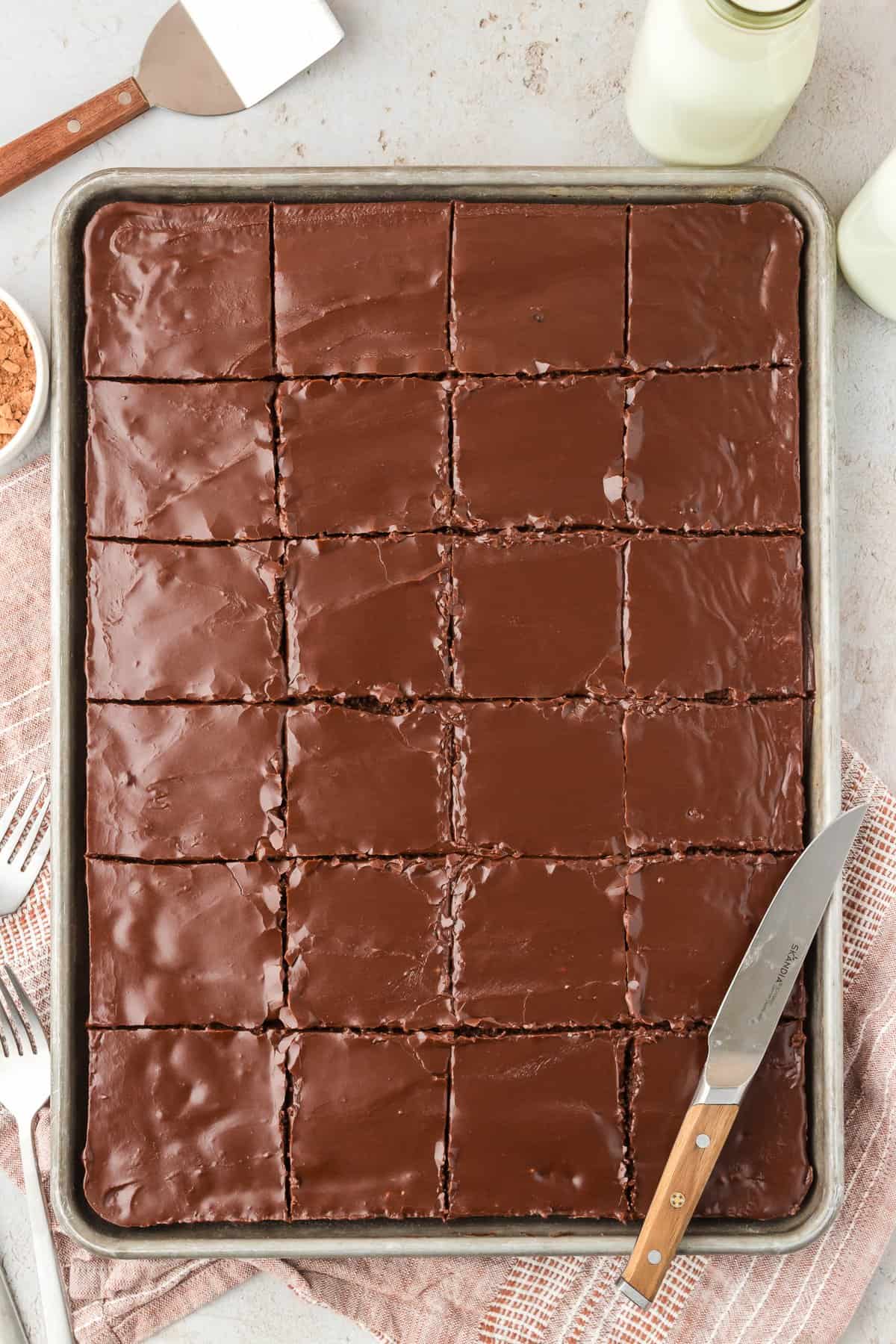 a sheet pan of texas sheet cake cut into squares with a knife resting on it, a spatula, milk, bowl of cocoa powder, fork, and tan kitchen towel around it