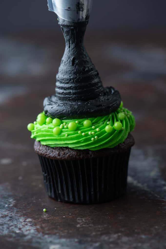 a chocolate cupcake with bright green frosting being piped with a circle of black frosting on top to shape a pointed witch hat