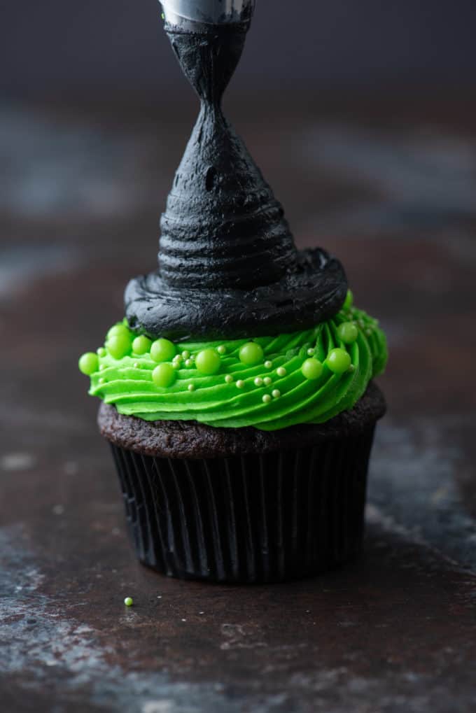 a chocolate cupcake with bright green frosting being piped with a circle of black frosting on top to shape a pointed witch hat