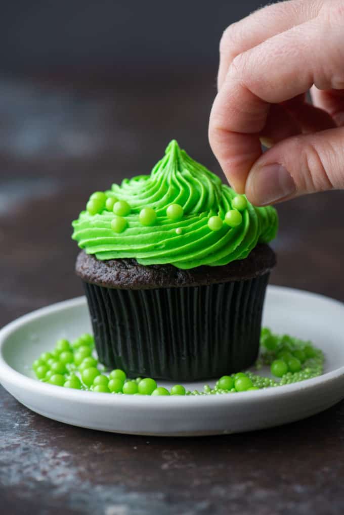 a chocolate cupcake with bright green frosting on a white plate being decorated being round green sprinkles and more sprinkles on the plate
