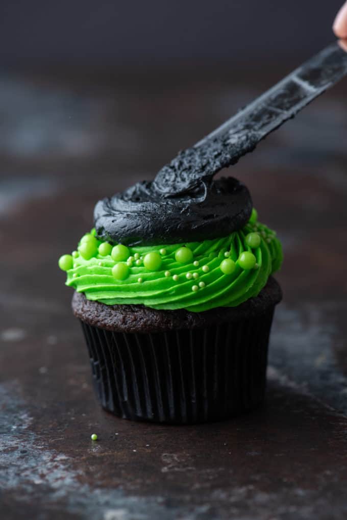 a chocolate cupcake with bright green frosting having black frosting on top being spread by a knife