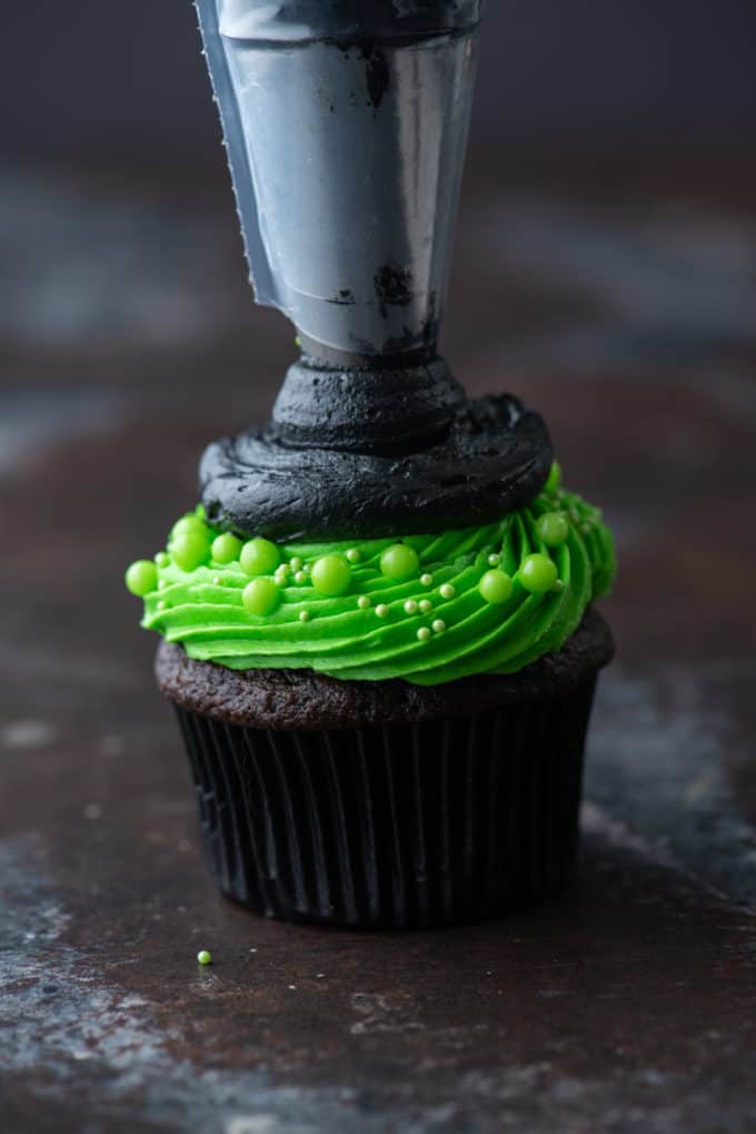 a chocolate cupcake with bright green frosting being piped with a circle of black frosting on top