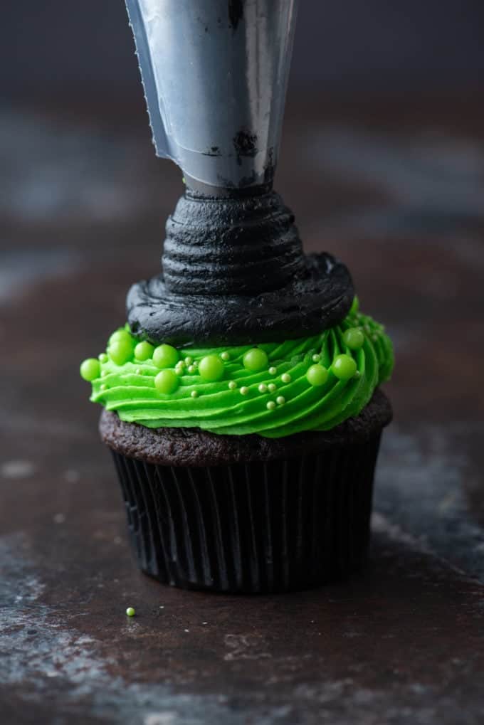 a chocolate cupcake with bright green frosting being piped with a circle of black frosting on top