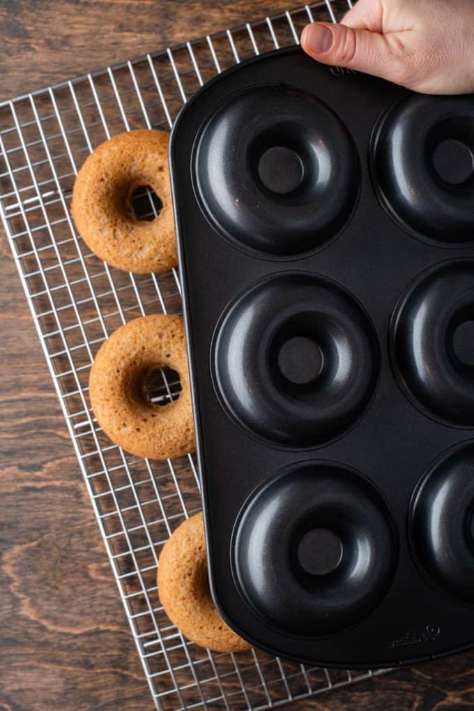 a donut pan flipped upside down being held over top of a wire rack with apple cider donuts on it on a wooden surface