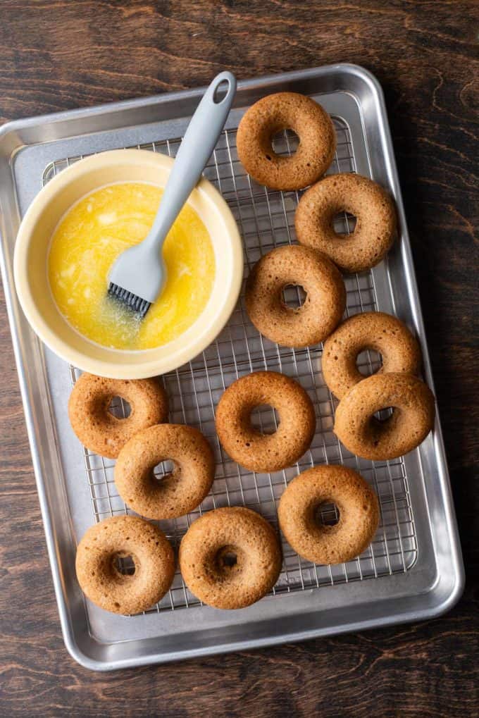 apple cider donuts on a wire rack in a sheet pan with a bowl of melted butter also on the wire rack with a basting brush in it