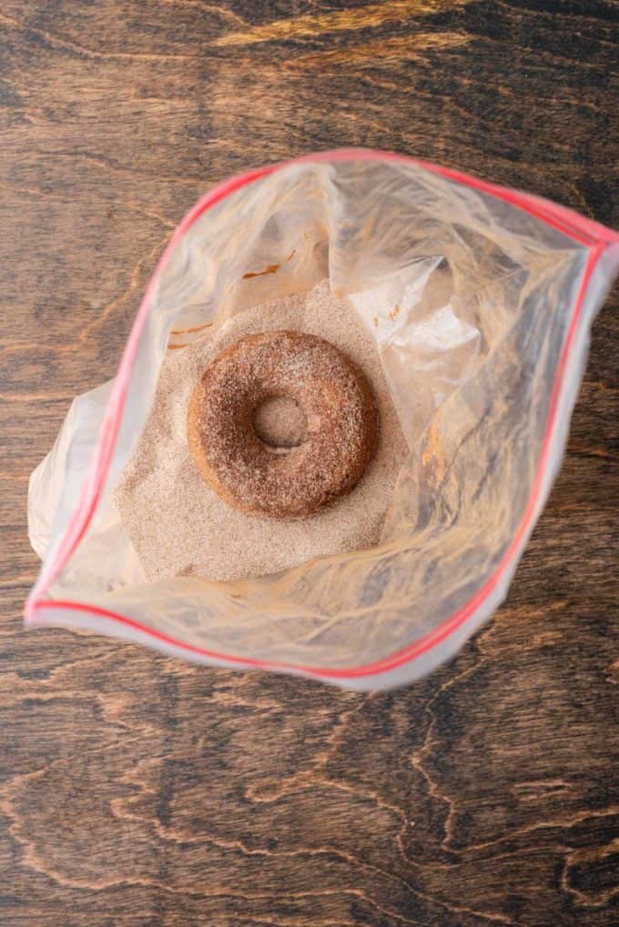 a baked apple cider donut inside a ziploc bag coated with a cinnamon sugar mixture on top of a wooden surface