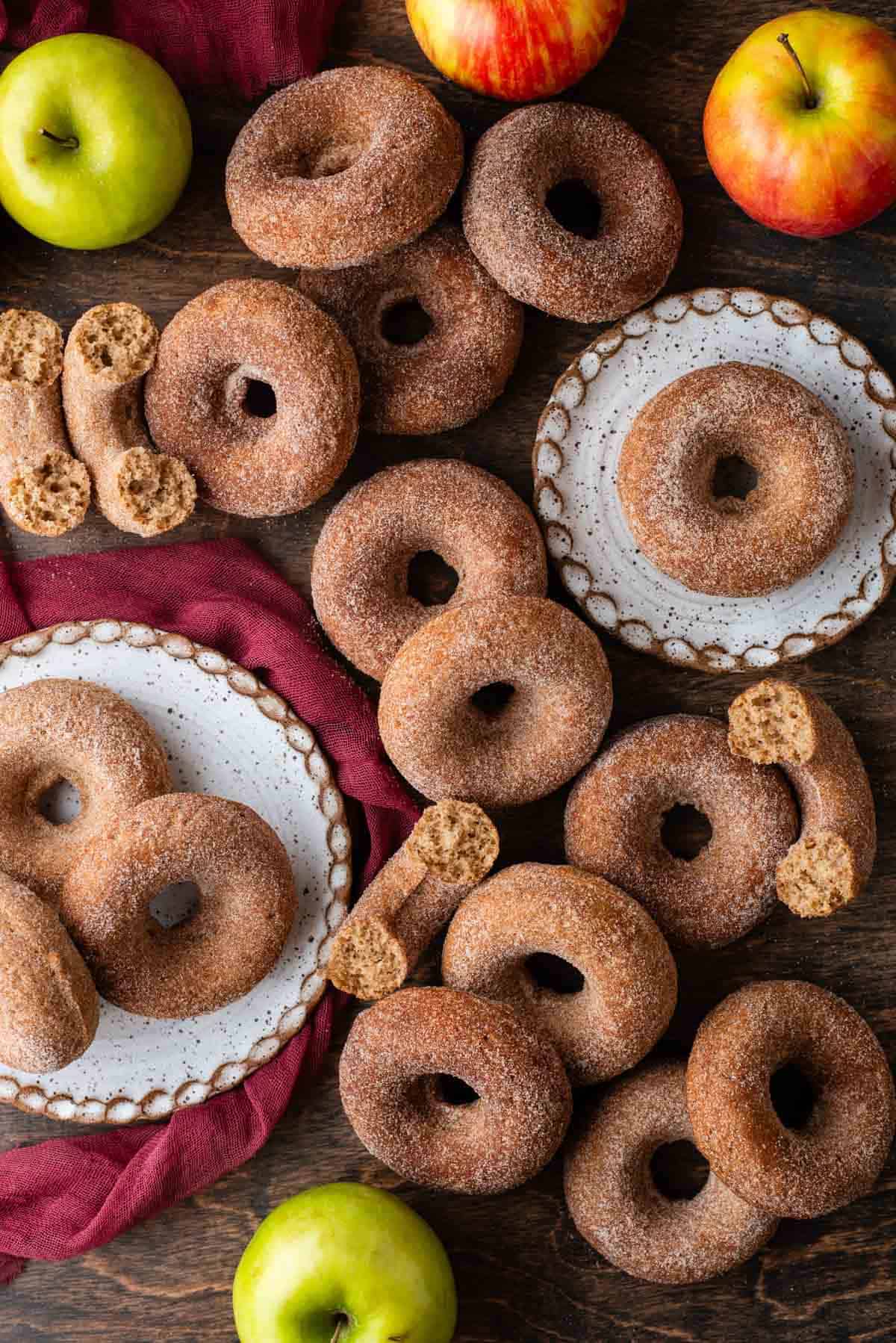apple cider donuts scattered around on a wooden surface, some on top different white plates, some in halves and some whole, with whole apples and a maroon towel