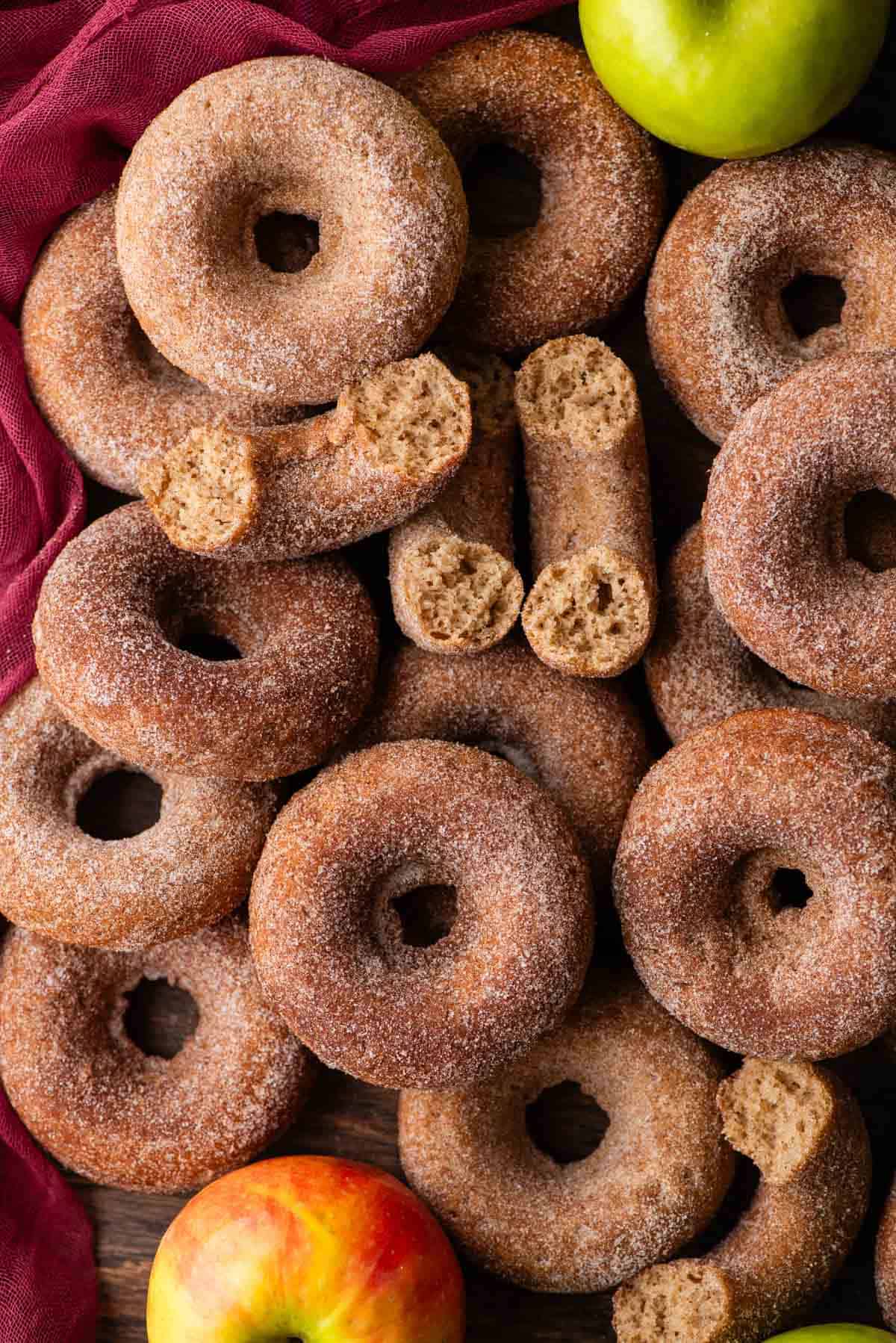a pile of apple cider donuts on a maroon towel, with a few half donuts on top and a whole green apple near the top right