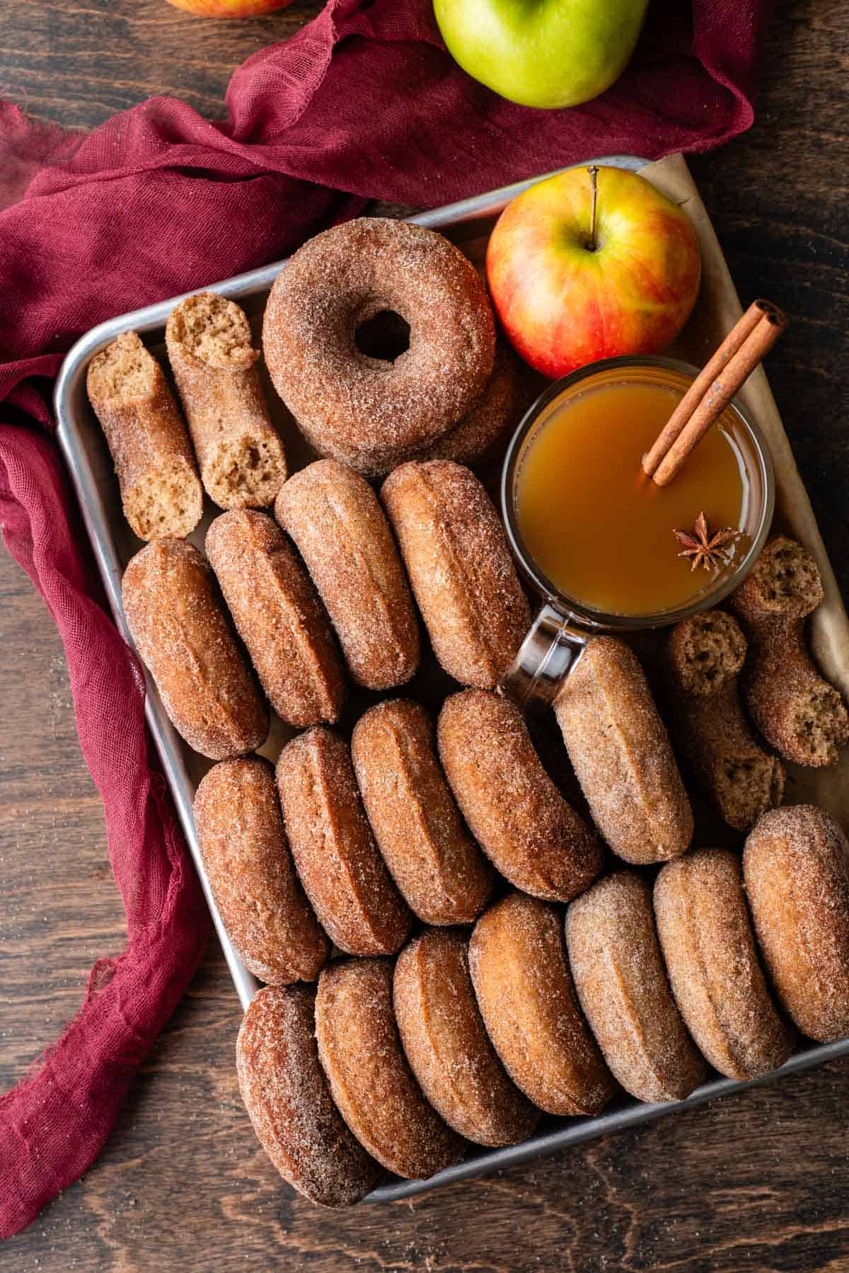 a sheet pan full of apple cider donuts, some lined up on their sides, some stacked and some in halves, with a glass of apple cider with a cinnamon stick in it, and a red and yellow apple, on top of a maroon towel with a whole green apple on it