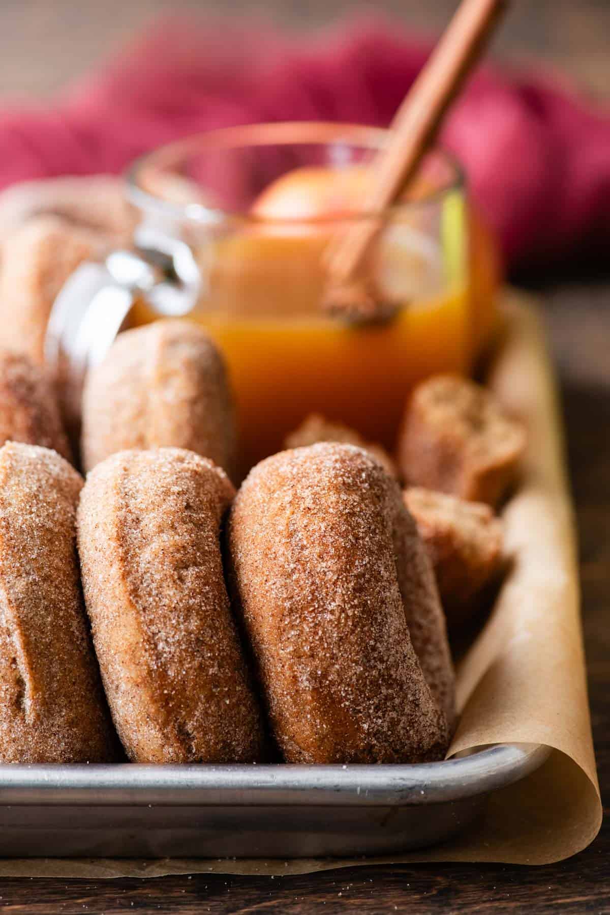 a sheet pan lined with parchment paper with apple cider donuts lined up on their sides in it and a cup of apple cider in the background