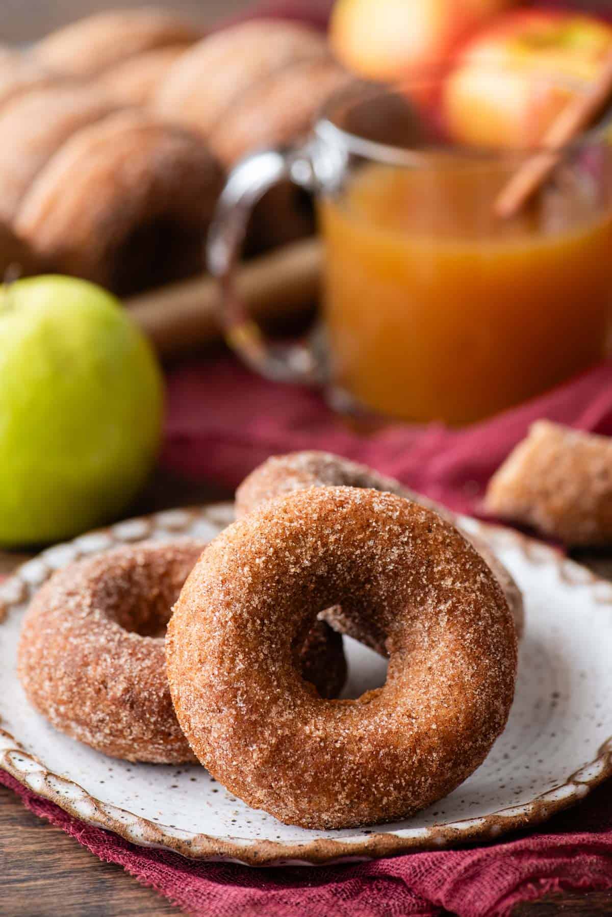three apple cider donuts on a plate that is sitting on top of a maroon kitchen towel, surrounded by a whole green apple, a cup of apple cider, and more donuts