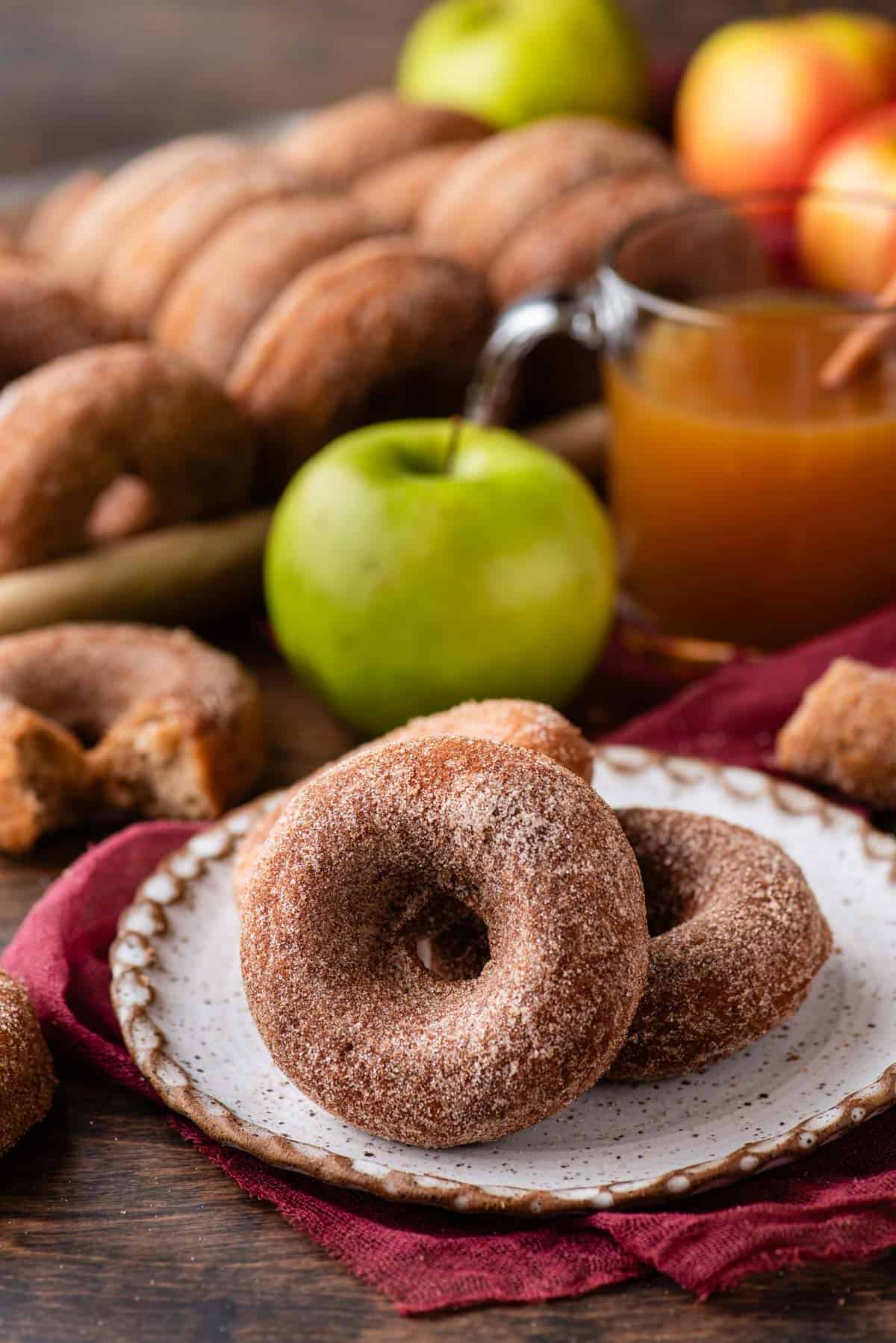 two apple cider donuts on a plate that is sitting on top of a maroon kitchen towel, surrounded by a whole green apple, a cup of apple cider, and more donuts