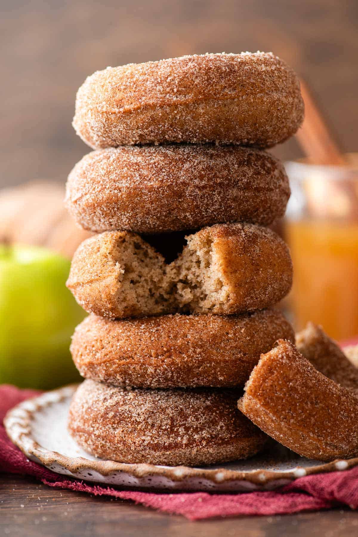 a stack of apple cider donuts with half a donut leaning on the side of it and the middle donut with a bite out, on a plate on top of a maroon towel, with a green apple and cup of apple cider in the background