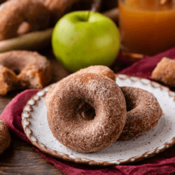 two apple cider donuts on a plate that is sitting on top of a maroon kitchen towel, surrounded by a whole green apple, a cup of apple cider, and more donuts