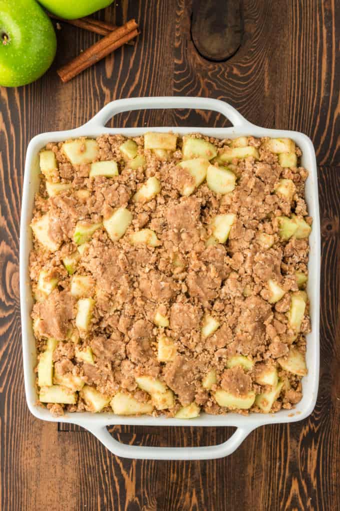 a white baking dish with apple crumble that is ready to bake in it on a wood surface surrounded by green apples and cinnamon sticks