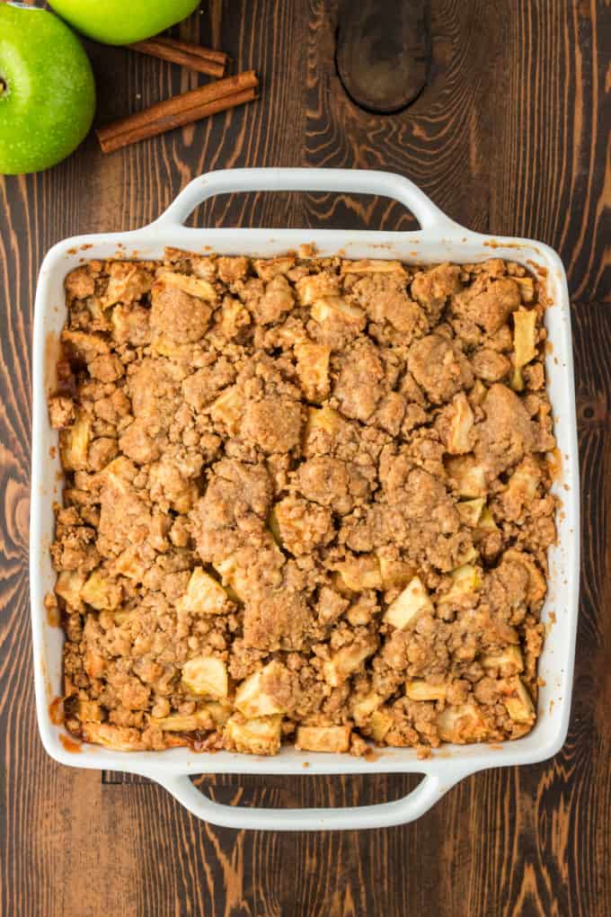 a white baking dish with apple crumble that is freshly baked in it on a wood surface surrounded by green apples and cinnamon sticks