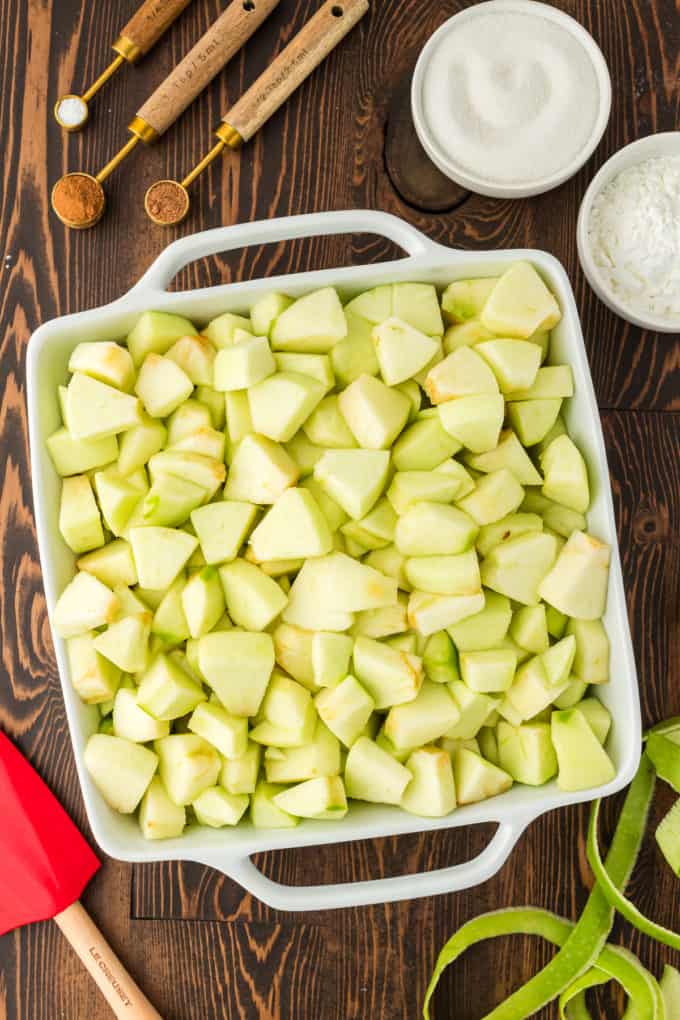 a white baking dish full of chopped green apples on a wood surface surrounded by measuring spoons and small bowls full of sugar, cornstarch, cinnamon, and nutmeg, a red spatula and green apple peels
