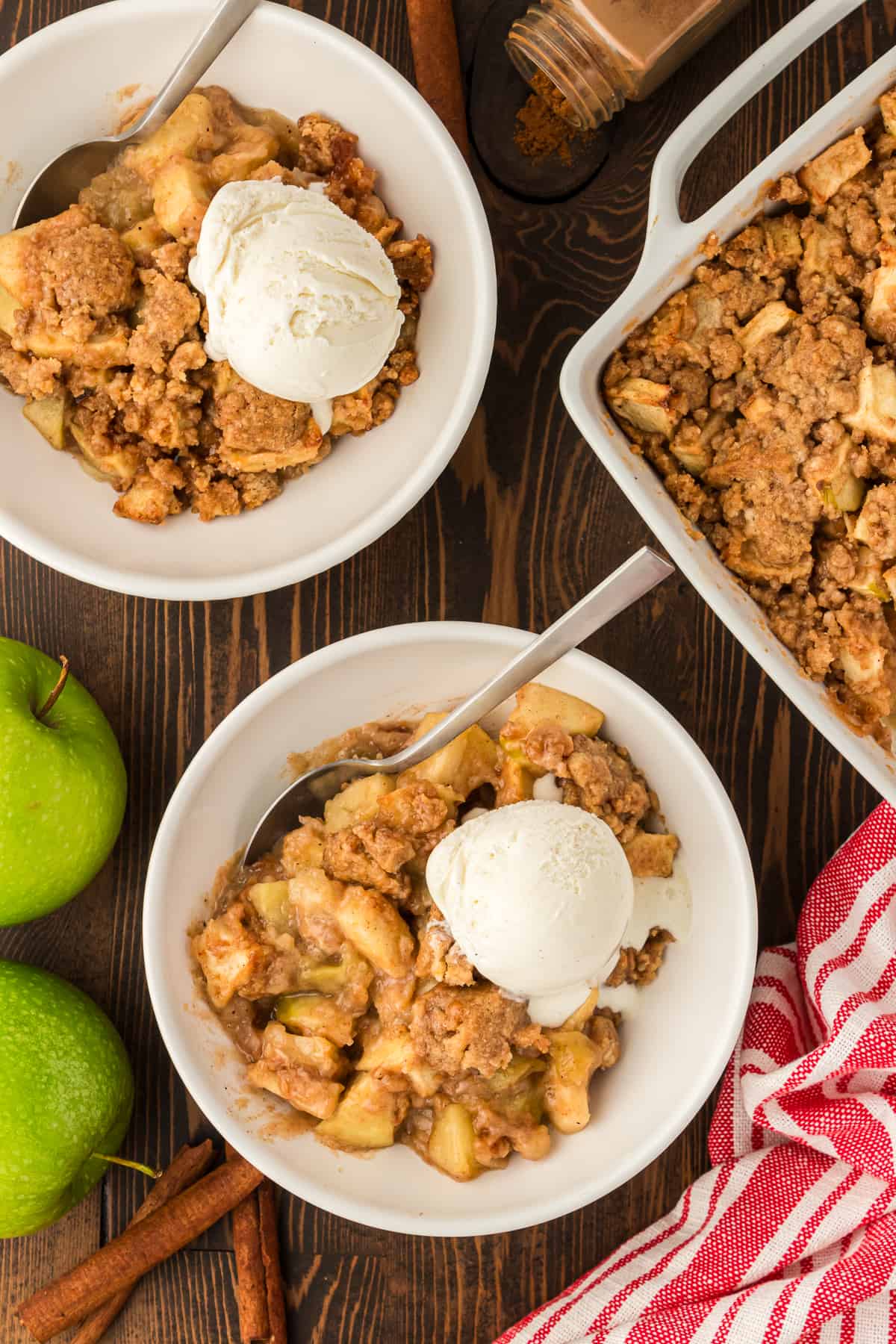 two bowls of apple crumble in a white bowl with a scoop of vanilla ice cream and a spoon, beside a white baking dish full of apple crumble, all on a wooden surface surrounded by a red and white striped kitchen towel, two green apples, cinnamon sticks and some spices