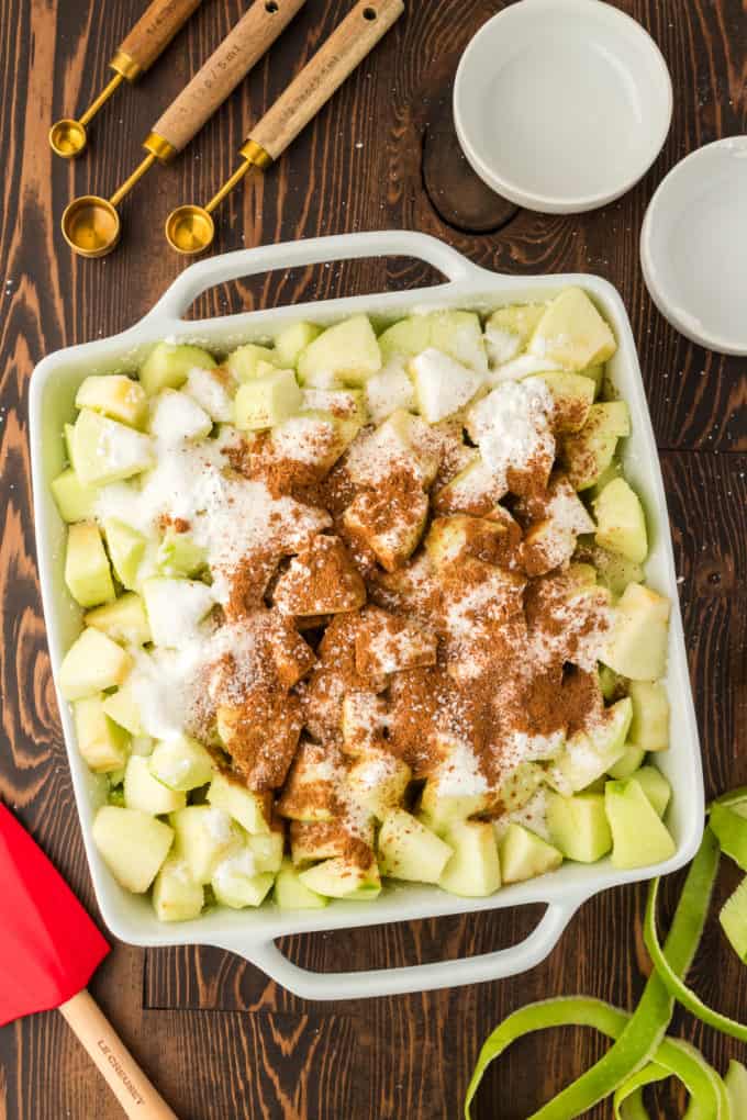a white baking dish full of chopped green apples on a wood surface topped with sugar, cornstarch, cinnamon, and nutmeg, surrounded by empty measuring spoons and bowls, a red spatula and green apple peels