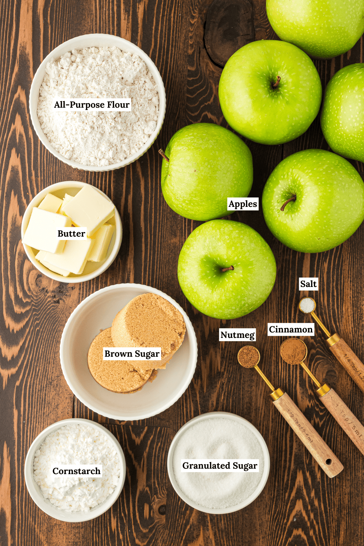 ingredients for apple crumble arranged around each other on a wooden surface including green apples, all-purpose flour, butter, brown sugar, salt, cinnamon, nutmeg, granulated sugar, and cornstarch
